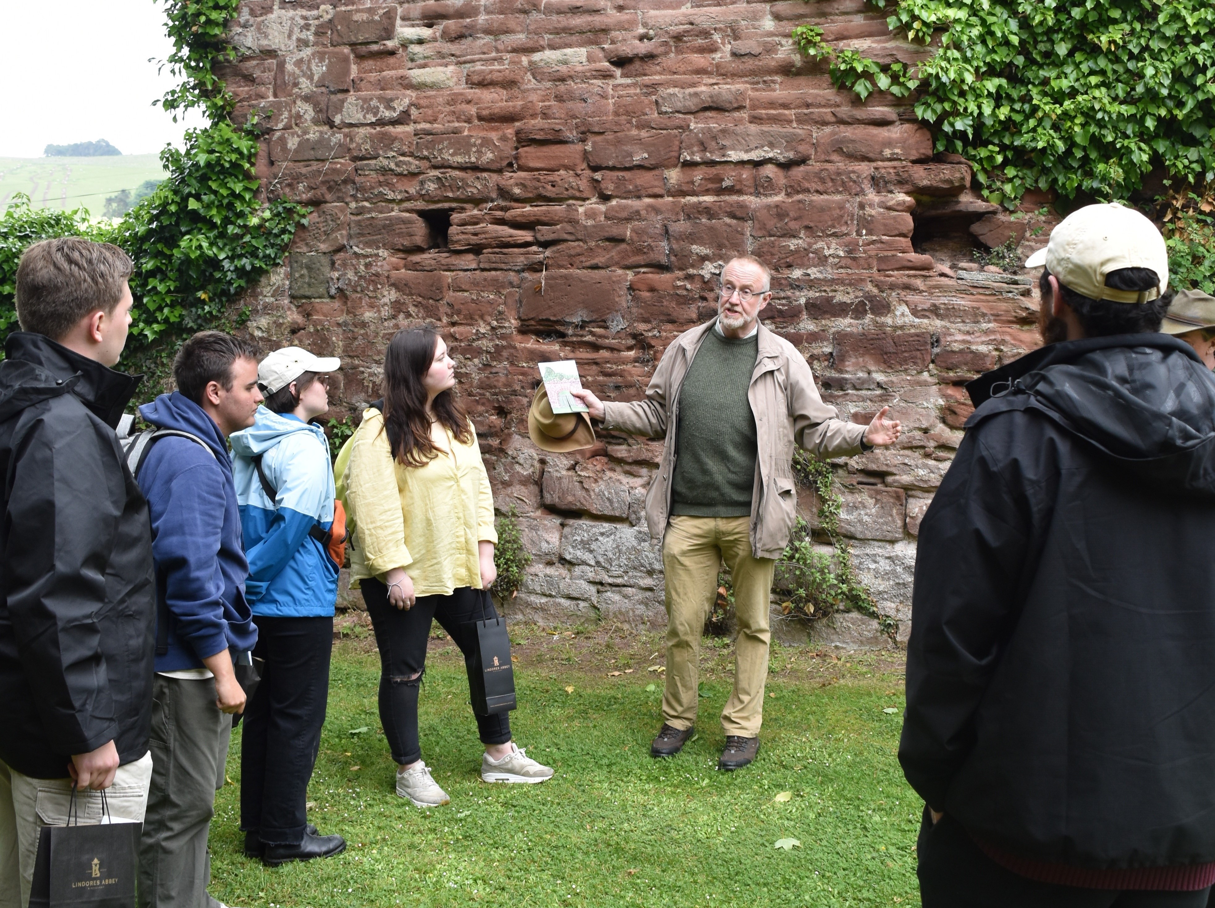 A lecturer speaks to group of students outoors, in front of the wall of a medieval structure.